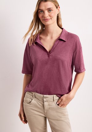 Mujer con una camisa de color malva de manga corta y cuello, y pantalones beige, de pie con una mano en el bolsillo sobre un fondo claro y liso.