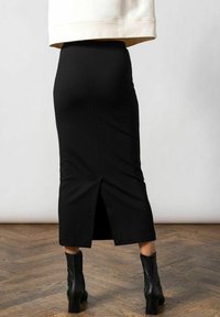 Sort pencilskør med en slids bagpå, lavet af strækbart stof, tætsiddende design, der når under knæet, parret med sorte ankelstøvler.