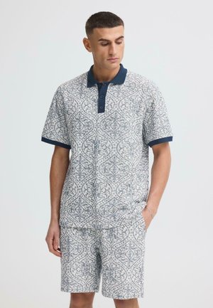 Mann trägt kurzärmeliges Poloshirt mit Muster, marineblauem Kragen und passenden gemusterten Shorts, steht mit einer Hand in der Tasche.