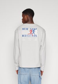 Lys grå langermet T-skjorte med et fargerikt New York Marathon-trykk på ryggen, med løpefigurer i rødt, blått og hvitt.