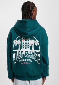 Tölgyzöld kapucnis pulóver, amelynek hátán egy nagy fehér grafika látható "JUST RHYSE" és "SUNSET MOTEL" felirattal, pálmafákkal és egy épület mintázattal.