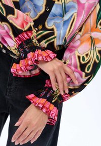 Ręce spoczywają na czarnych spodniach, nosząc kolorową bluzkę z długim rękawem, z elastycznymi, falbankowymi mankietami i wzorem kwiatowym w odcieniach różu, niebieskiego i pomarańczowego.