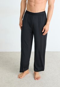 Homme portant un pantalon de pyjama noir amples, debout pieds nus sur un tapis beige contre un mur blanc.