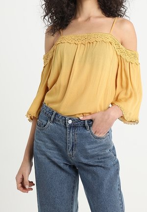 Off-the-shoulder gele top met kantafwerking, losse pasvorm, korte geflardeerde mouwen, aangekleed met hoogwaisted blauwe denim jeans.