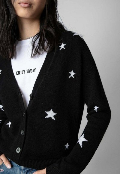 Cardigan noir avec des motifs d'étoiles blanches, comportant un col en V, une fermeture à boutons et des poignets côtelés. Porté par-dessus une chemise blanche avec du texte.