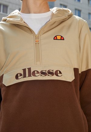 Personne portant un hoodie ellesse beige et marron avec une demi-fermeture éclair et un logo sur la poitrine, en extérieur avec une chemise blanche en dessous.