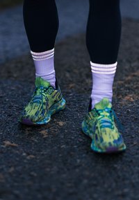 Groene en blauwe sportieve schoenen met een gepattern surface, platte veters en paarse accenten, gecombineerd met witte en lila gestreepte sokken.