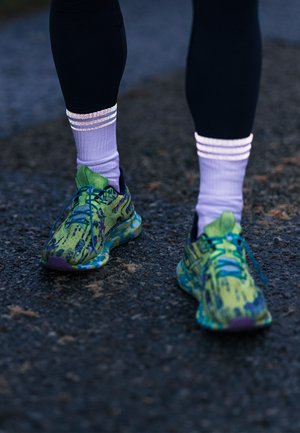 Grüne und blaue gemusterte Sportschuhe mit einer strukturierten Oberfläche, flachen Schnürsenkeln und lila Akzenten, kombiniert mit weißen und lila gestreiften Socken.