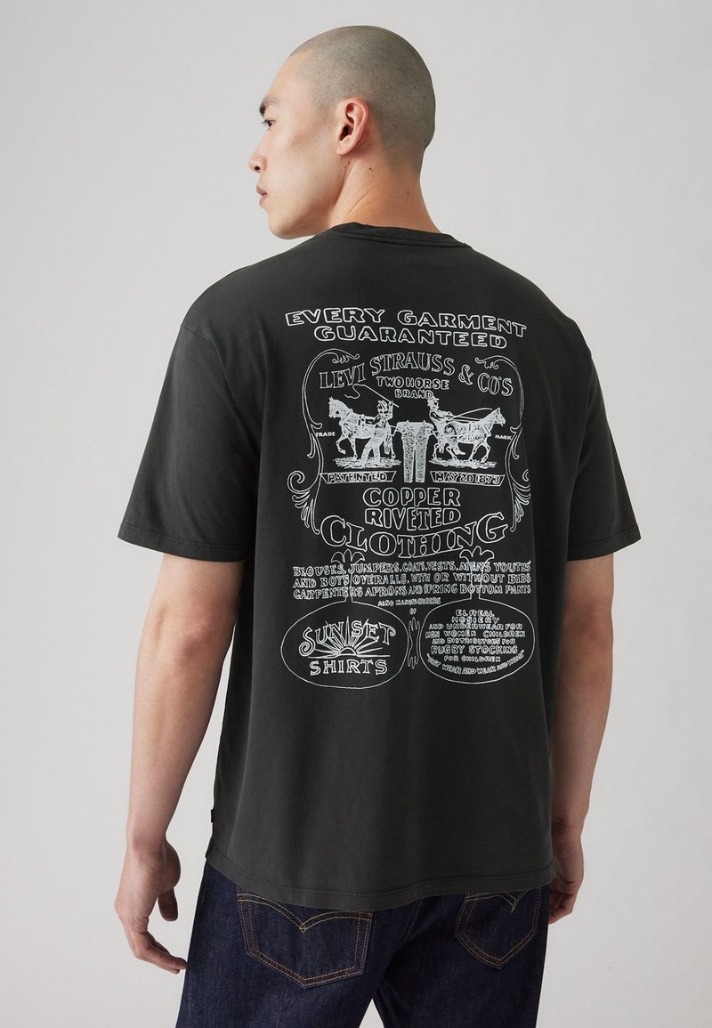 La camiseta de algodón negra presenta una impresión gráfica en la parte posterior, mostrando tipografía vintage y diseños relacionados con Levi Strauss & Co.