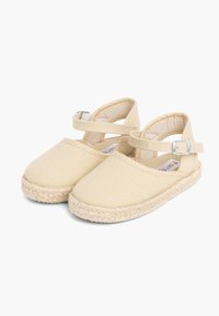 Espadrilles en toile beige avec une semelle en jute tissée, présentées dans un design à bout fermé et dotées d'une sangle chevillère ajustable avec une boucle métallique.