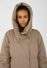 Veste rembourrée marron avec une grande capuche enveloppante, des accents de boutons et une texture douce, dotée d'un col montant pour plus de chaleur.