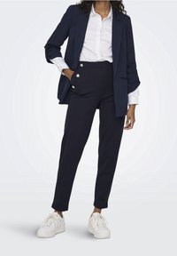 Blazer marine avec pantalon taille haute assorti orné de boutons dorés, associé à une chemise blanche et des baskets blanches épaisses.