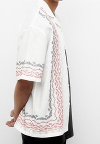 Kortærmet, hvidt linnedskjorte med intrikate røde og sorte broderier langs kanterne og knapper ned foran.