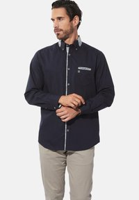 Marineblaue Button-Up-Hemd mit gemustertem Kragen und Taschenbesatz, mit langen Ärmeln und tailliertem Schnitt. Die Knöpfe sind weiß, der Stoff ist glatt.