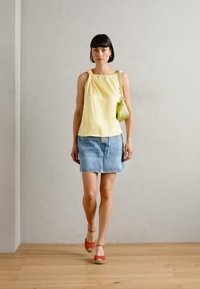 Gelbes ärmelloses Oberteil mit geraffter Detail, kombiniert mit einem hellblauen Jeansrock und roten Espadrilles. Eine grüne Tasche hängt über der Schulter.