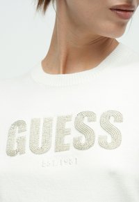 Λευκό πλεκτό πουλόβερ με τη λέξη "GUESS" από στρας και το κέντημα "EST.1981" από κάτω. Υφή υφάσματος με ριμπ λαιμόκοψη.