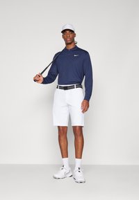 Marinblå långärmad polo, vita shorts, svart och vit skärp, och vita sportskor. Håller golfklubba. Enkel bakgrund.