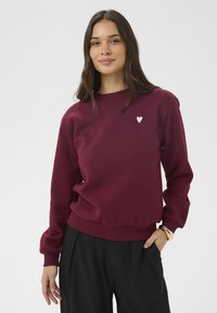 Burgundská mikina s žebrovaným kulatým výstřihem, elastickým pasem a malým bílým srdíčkovým logem na levé straně hrudi. Hladká textura.