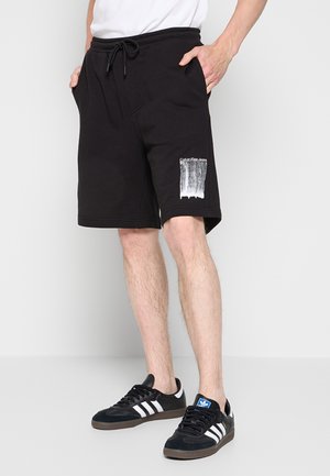 Uomo che indossa pantaloncini neri Calvin Klein con stampa bianca sulla gamba sinistra, sneakers nere Adidas con strisce bianche e una camicia bianca.