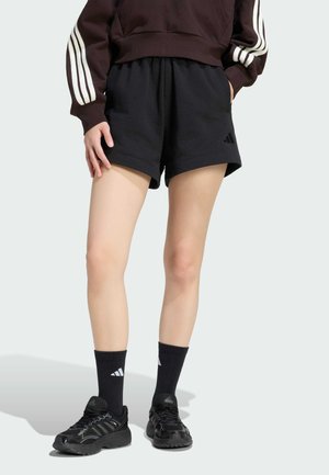 Personne portant un short de sport noir, des chaussettes noires montantes, des baskets noires, et un sweat sombre avec des bandes blanches sur les manches.