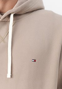 Beżowa bluza z kapturem z regulowanym sznurkiem, z małą naszywką z logo w czerwonym, białym i niebieskim kolorze na lewej piersi. Miękki, fakturowany materiał.