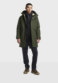 Olijfgroene parka met bont gevoerde capuchon, zwarte binnenjas met rits, donkere broek en bruine wandelschoenen. Voorzien van zakken en een comfortabele pasvorm.