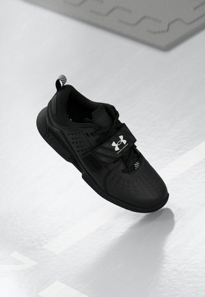 Chaussure de sport noire avec logo Under Armour, dotée de lacets et d'une sangle en velcro, présentée sur un fond clair et texturé.