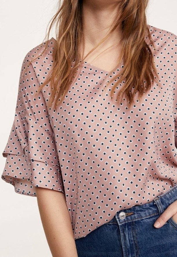Kvinde iført en mauve kortærmet bluse med lagdelte flæser og et lille sort prikmønster, parret med blå denim jeans.