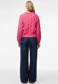 Blond persoon in een getextureerde roze gebreide trui en wijde donkerblauwe jeans, staand met de rug naar de camera tegen een witte achtergrond.