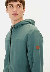 Veste à capuche en tissu teal, fermeture éclair à l'avant, avec un panneau sur la poitrine, une capuche réglable et un patch logo brun sur la manche gauche.