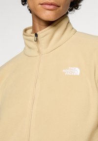 Közeli kép egy személyről, aki bézs színű cipzáras polárdzsekit visel álló gallérral és a mellkasán "The North Face" logóval.