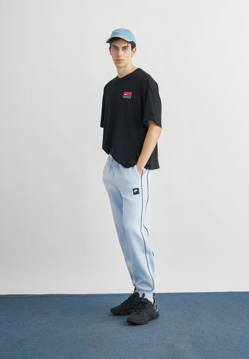 Giovane uomo con indosso una t-shirt nera Nike, pantaloni della tuta azzurri, scarpe da ginnastica nere e un cappellino azzurro, in piedi con le mani nelle tasche.