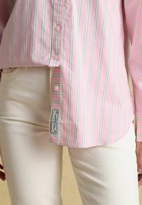 Chemise rose à boutons avec des fines rayures verticales vertes et blanches, portée non rentrée sur un pantalon blanc cassé, avec une étiquette de marque visible près de l'ourlet.