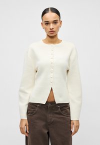 Balts adīts kardigans ar gariem piedurknēm, apaļu kakla izgriezumu un priekšējām pogām. Ir taisna apakšmala un brīvs siluets.