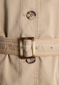 Beige stoffen riem met een grote rechthoekige bruine en crèmekleurige gesp, voorzien van knoopdetails en zichtbare stiksels. Smooth textuur door het geheel.