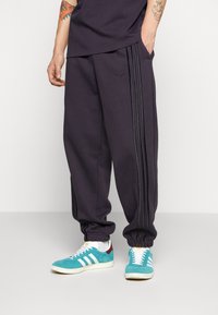 Pantalones de chándal morados oscuros con bolsillos laterales, puños elásticos y tres rayas negras a lo largo de los lados, combinados con zapatillas en tonos verde azulado y blanco.