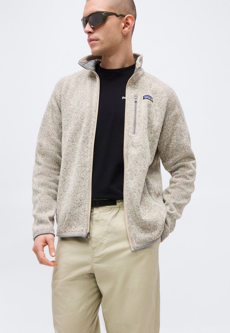 Man wearing beige zip-up fleece jacket, black t-shirt, light khaki pants, and dark sunglasses, looking to the side with one hand in pocket.