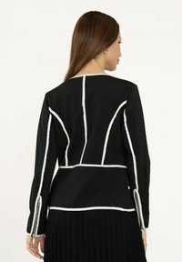 Veste noire avec accents de coutures blanches, forme structurée, manches longues et fermetures éclair aux poignets. Tissu lisse, design minimaliste.