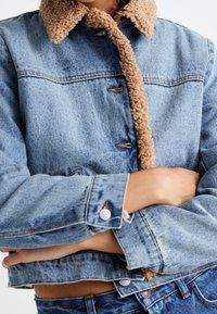 Džínová bunda s světlým modrým praním, hnědým beránkovým límcem, zapínáním na knoflíky a ohrnutými rukávy s kovovými knoflíky.
