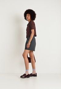 Καφέ υφασμάτινο μπλουζάκι κοντομάνικο σε συνδυασμό με γκρι φούστα. Το μοντέλο φοράει σκούρα παπούτσια και μωβ καλσόν με πουά. Μινιμαλιστικό φόντο.