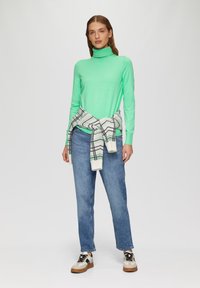 Mintgroene coltrui met lange mouwen, gecombineerd met lichtblauwe rechte spijkerbroek. Een grijs geruite trui om de taille geknoopt. Casual schoenen.