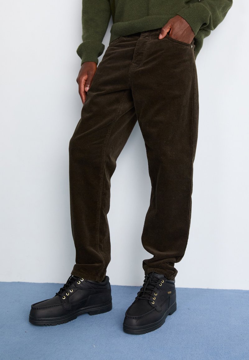 Braune Baumwollcord-Hosen mit geradem Bein, kombiniert mit schwarzen Lederschuhen mit goldenen Ösen und strukturierten Sohlen.