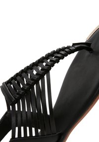 Black sandal with braided upper straps, flat sole, smooth leather texture, and contrasting tan outsole. Minimalist design with no visible hardware.