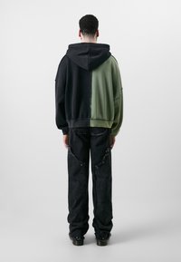 Hooded sweatshirt features black and olive green color blocks with raw edges, paired with black denim pants with distressed accents.