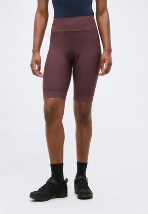 Personne portant un cuissard de cyclisme matelassé violet foncé, des chaussettes noires et des chaussures de cyclisme noires, debout devant un fond blanc uni.