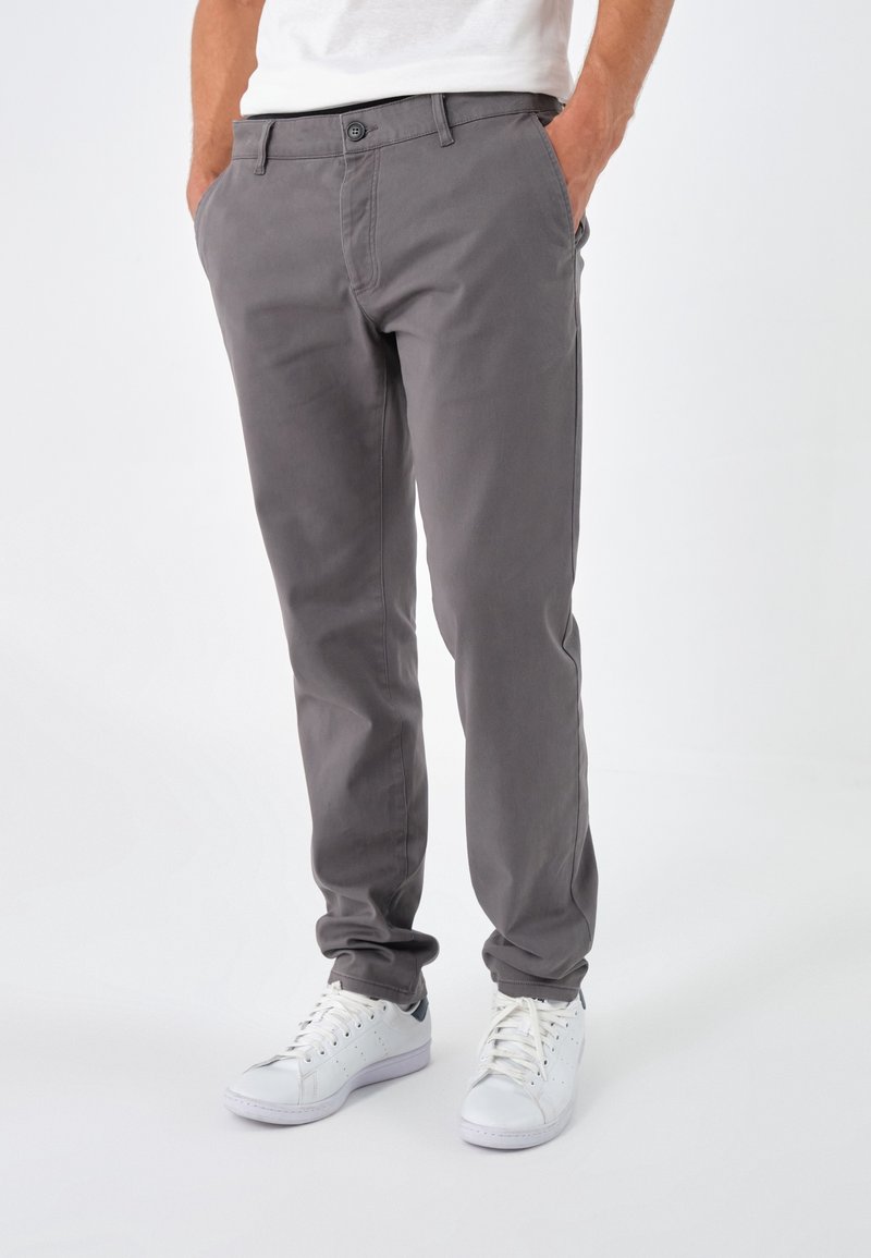 Homme portant un pantalon droit gris et des baskets blanches, debout les mains dans les poches devant un fond clair uni.