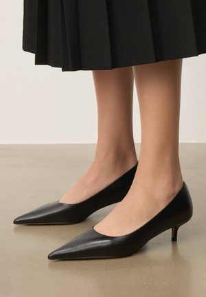 Noge v črnih usnjenih čevljih z ostrim sprednjim delom in nizkimi petami, združene s črno pliseasto krilo, stojijo na gladkem tleh.