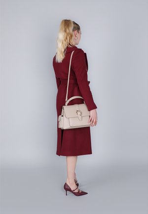 Beige leren handtas met een klep, gouden hardware en een afneembare schouderband. Draagt met een bordeauxrode jas en bijpassende hakken.