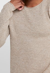 Närbild av en person som bär en beige, strukturerad stickad tröja, hand som rör vid halsen, vilket framhäver tygdetaljer och passform.