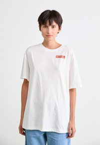 White cotton t-shirt with short sleeves and a round neckline. Features a small red logo on the left chest area. Loose-fitting design.